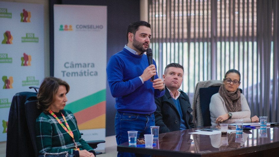 Gabriel em pé, atrás de uma mesa falando ao microfone e sendo olhado por mais três pessoas que compõem a mesa junto com ele.