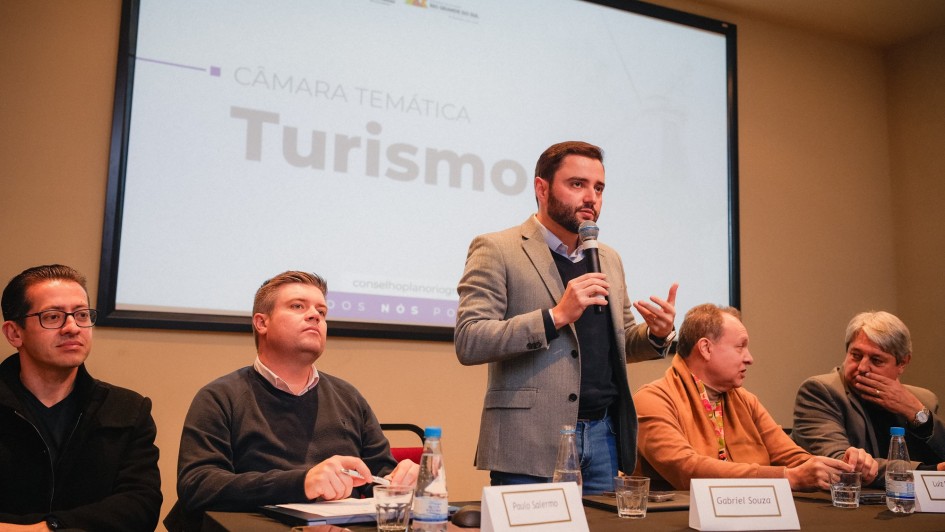 Reunião da Câmara Temática do Turismo em que Gabriel está ao microfone falando em pé atrás de uma bancada. Ao lado dele, dois homens estão sentados à esquerda e dois homens estão sentados à direita.